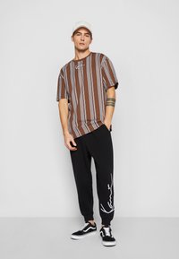 Brun kortermet skjorte med vertikale blå striper, svarte joggebukser med hvit grafisk design, og svarte sneakers med hvite såler.