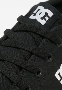Black canvas sneaker featuring a textured surface, flat black laces, metal eyelets, and a white logo patch on the tongue.
