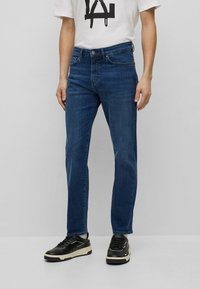 Dark blue denim jeans with a straight leg design, five pockets, and a button-fly closure. Worn with a white graphic t-shirt and black sneakers.