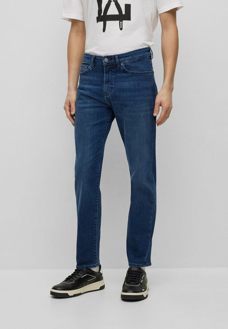 Dark blue denim jeans with a straight leg design, five pockets, and a button-fly closure. Worn with a white graphic t-shirt and black sneakers.