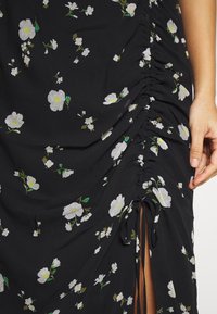 Černé květinové šaty s bílými květy a zelenými akcenty. Disponují nařasenou strukturou a stuhami po straně pro nastavitelný střih.