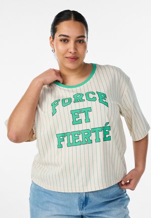 Kobieta o ciemnych włosach zaczesanych do tyłu, ubrana w kremową koszulę z zielonymi paskami i napisem "FORCE ET FIERTÉ" oraz jasnoniebieskie dżinsy.