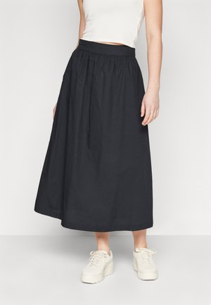 Femme portant une jupe midi noire avec taille froncée, un top court blanc et des baskets compensées blanches, debout devant un fond blanc uni.