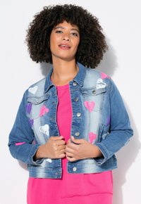 Chaqueta de mezclilla con patrones de corazones en rosa, púrpura y blanco, cuello clásico, bolsillos frontales y botones metálicos. Usada sobre una camiseta rosa.