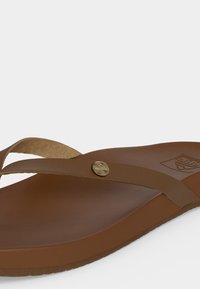 Καφέ δερμάτινο σανδάλι τύπου φλιπ-φλοπ με απλό λουρί που διασχίζει το πόδι και ένα μικρό μεταλλικό κουμπί με λογότυπο στο λουρί.