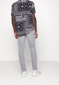 Camicia nera a maniche corte con motivi paisley e puntini bianchi, abbinata a pantaloni grigio chiaro slim fit e scarpe da ginnastica grigie.