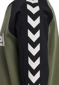 Schwarzer Ärmel mit weißen nach unten zeigenden Chevron-Pfeilen auf einem olivgrünen Kleidungsstück.