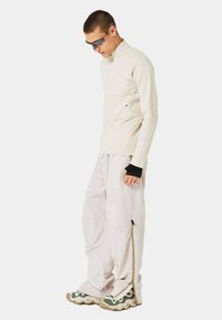 Helles beige Langarmoberteil mit strukturiertem Design, kombiniert mit lockeren, cremefarbenen Hosen mit seitlichen Reißverschlüssen und Schuhwerk mit grünen Akzenten.
