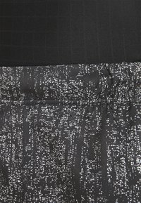 Schwarzes Sporttop mit einem strukturierten Gittermuster, kombiniert mit einem schwarz-weißen Stoff mit unregelmäßigen Linien und einem elastischen Bund.