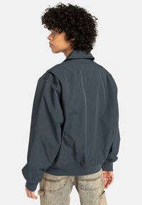 Person with curly hair wearing a dark gray jacket and light-colored denim cargo pants, facing sideways with a neutral background.