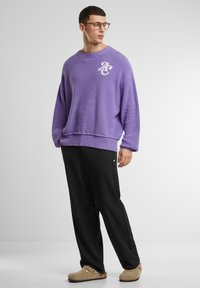 Lila Waffelstrick-Pullover mit einem gerippten Halsausschnitt, kombiniert mit schwarzen Hosen. Das Outfit wird mit beigefarbenen Slippern abgerundet.