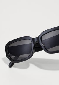 Black oversized sunglasses with a geometric design, featuring dark lenses and a matte finish on the frame. Distinctive angular shape.