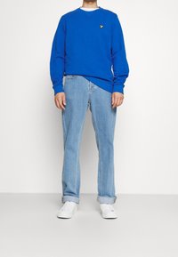 Mörkblå herr sweatshirt med en gul logga, kombinerad med ljusblå jeans och vita sneakers. Tröjan har rundad halsringning och ribbade ärmslut.