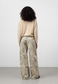 Osoba z kręconymi włosami, ubrana w beżowy, puszysty sweter i szerokie, marmurkowe spodnie, stoi przy gładkiej ścianie.