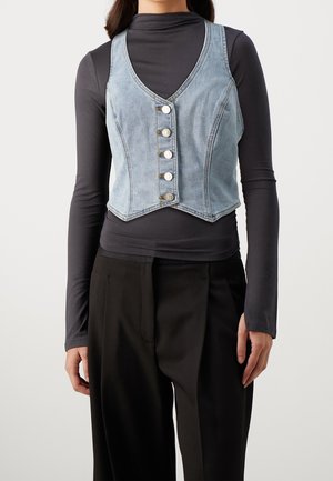 Femme portant un gilet en denim bleu clair boutonné par-dessus un top noir cintré à manches longues et un pantalon noir ample.