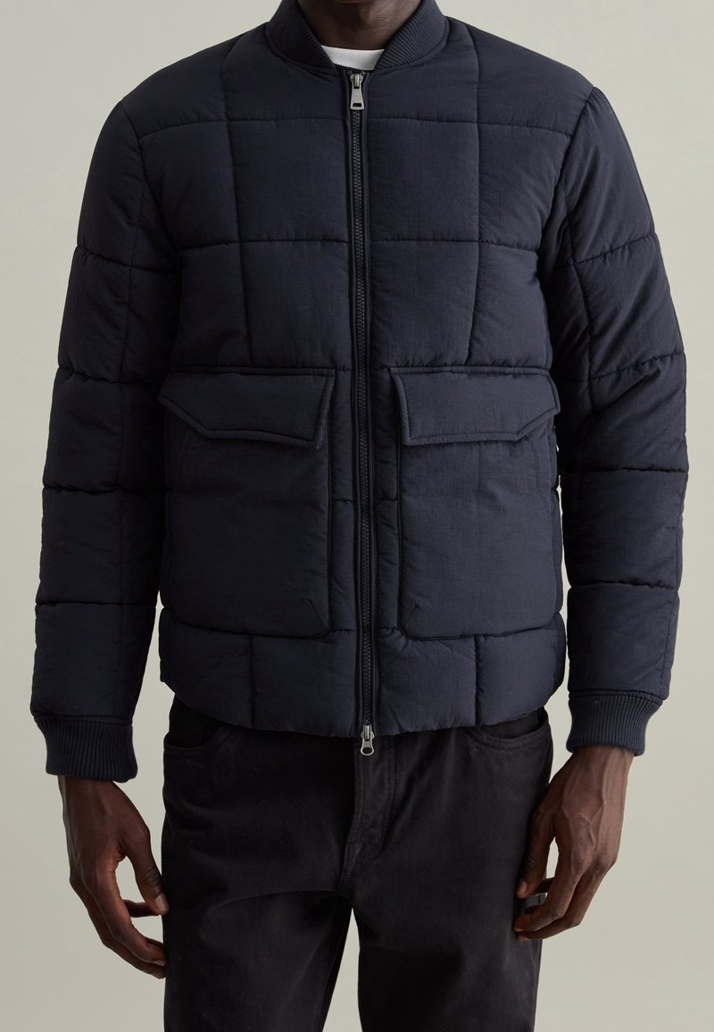 Giacca imbottita blu navy con design trapuntato, due tasche applicate anteriori, polsini a costine e chiusura con zip. Realizzata in tessuto leggero e strutturato.