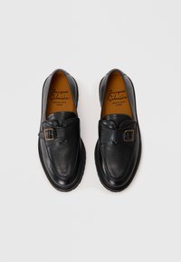 Black leather loafers with a textured finish, featuring a buckle accent, rounded toe, and a thick rubber sole. Interior has a tan color.