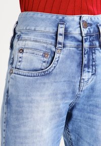 Vybledé světle modré džíny s přiléhavým střihem, které mají přední kapsy, kovový knoflík a švy s kontrastním prošitím.