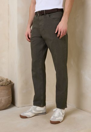 Mann trägt dunkelgrüne Hose, braunen Gürtel, weiße und graue Turnschuhe und steht auf einem hellfarbenen Boden in der Nähe eines dekorativen Topfes.
