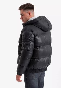 Zwarte gewatteerde jas met een capuchon, voorzien van een ritssluiting, elastische manchetten en een patch op de linker mouw. Glad, glanzend oppervlak.
