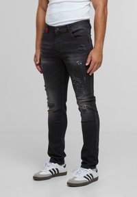 Mees, kellel on seljas mustad kitsad kulunud teksad ja valged tossud mustade triipudega, seisab ühtlasel hallil taustal.