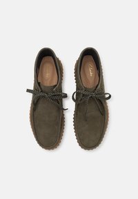 Grüne Wildlederschuhe mit Gummisohlen, die eine strukturierte Oberfläche und kontrastierende schwarz-weiße Schnürsenkel aufweisen. Clarks-Logo innen.