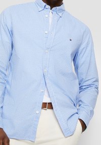 Camicia button-up a quadretti azzurri chiari con colletto, dotata di bottoni bianchi e un piccolo logo sul petto, abbinata a una maglietta bianca sotto.