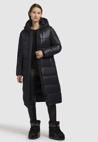 Langer schwarzer Daunenmantel mit Kapuze, ausgestattet mit einem gesteppten Design, Frontreißverschluss und Seitentaschen, kombiniert mit glänzenden schwarzen Stiefeletten.