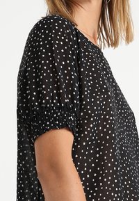Černá blůza s bílým rozptýleným tečkovaným vzorem, krátké nabírané rukávy s elastickými manžetami a lehkým, průsvitným textilním vzhledem.