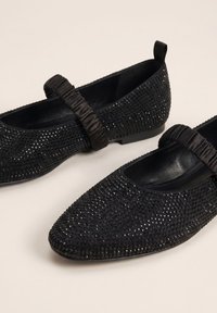 Schwarze flache Schuhe mit spitzen Zehen, verziert mit kleinen glänzenden Nieten und elastischen gerafften Riemen über dem Spann.