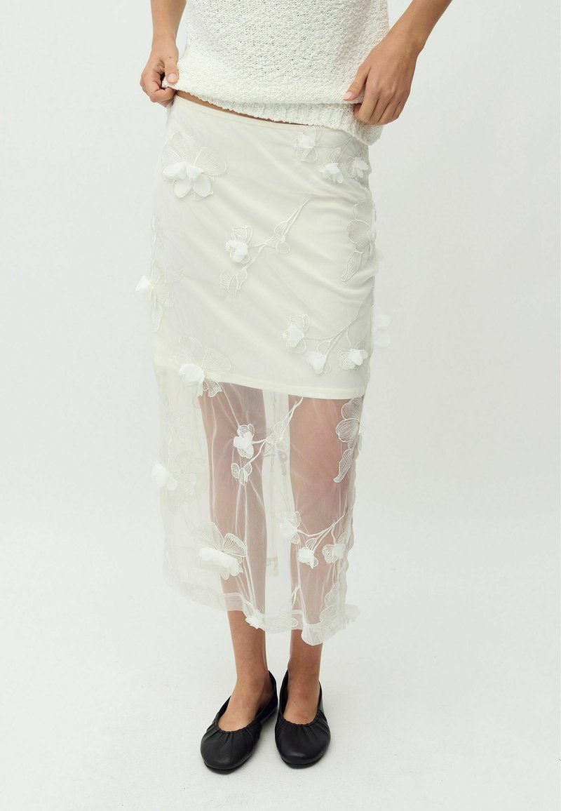 Falda blanca transparente con bordado floral, superpuesta sobre una base sólida. Presenta una cintura ajustada y cae hasta la mitad de la pantorrilla. Se lleva con zapatos planos negros.