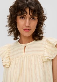 Vrouw met krullend bruin haar, gekleed in een lichtgele gestreepte blouse met ruches, kijkt recht in de camera.