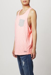 Ljusrosa linne med en grå ficka; lös passform, rundad halsringning och sidofickor. Bärs med slitna blå jeans.