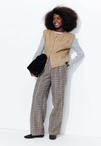 Gilet en laine beige avec des boutons dorés sur une chemise à rayures à manches longues, associé à un pantalon large à carreaux et des ballerines noires à texture. Sac à main noir.