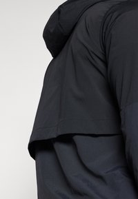 Black jacket with a hood, featuring a layered design and textured fabric. The hem is slightly longer at the back, creating contrast and depth.