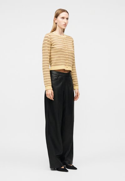 Γυναίκα που φοράει ένα κοντό μπεζ και καφετί ριγέ ζακέτα, φαρδιά μαύρη παντελόνια και μαύρα μυτερά παπούτσια, που στέκεται μπροστά σε απλό φόντο.