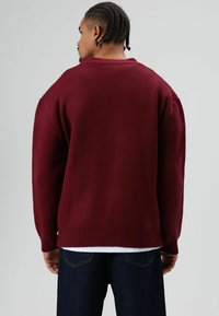Pull oversize bordeaux avec ourlet et poignets côtelés, confectionné dans un tissu doux, vu de dos. Assorti à un jean foncé.