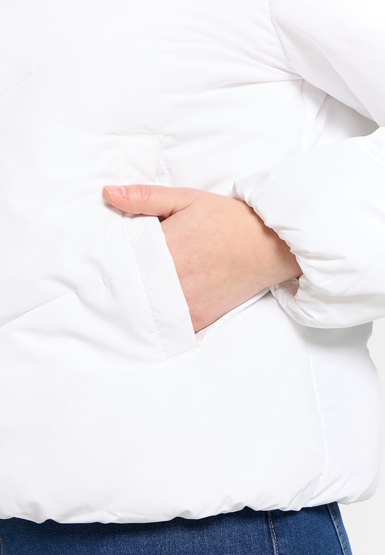 Hand in de zijzak van een wit gewatteerd winterjack met elastische manchetten, gedragen over een spijkerbroek.