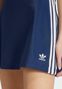 Sötétkék sport szoknya fehér oldalsávokkal, sima anyagból készült. Kis fehér Adidas logó található az alján.