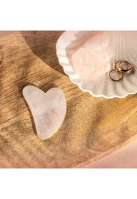 Heart-shaped rose quartz stone with a smooth surface, placed on a wooden tray next to a white shell dish containing pink crystals and rings.