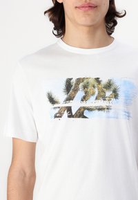 Hvid bomulds-T-shirt med et grafisk print af palmer mod en blå baggrund, med blød tekstur og korte ærmer.