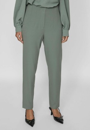 Pantalons ajustés vert clair avec des plis, une texture lisse et une coupe ajustée, associés à des talons aiguilles noirs.