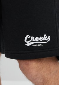Pantalones cortos negros de algodón con una textura suave. Presentan un logotipo blanco "Creeks Original" en el lateral, mostrando un diseño casual.