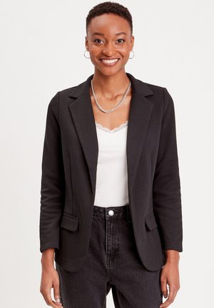Blazer nero con un design sartoriale, due tasche frontali e una texture liscia. Indossato sopra una maglietta bianca, abbinato a jeans scuri.