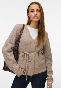 Svijetlosmeđi pleteni kardigan s V-izrezom, remenom oko struka i dugim rukavima. Nošen preko bijele košulje, u kombinaciji s tamnim trapericama i ispletenom torbom.