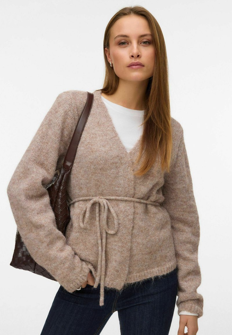 Svijetlosmeđi pleteni kardigan s V-izrezom, remenom oko struka i dugim rukavima. Nošen preko bijele košulje, u kombinaciji s tamnim trapericama i ispletenom torbom.