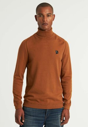 Pull col roulé rouille en tissu doux, offrant une coupe ajustée et des poignets côtelés, avec un petit logo noir sur la poitrine. Associé à un jean bleu.