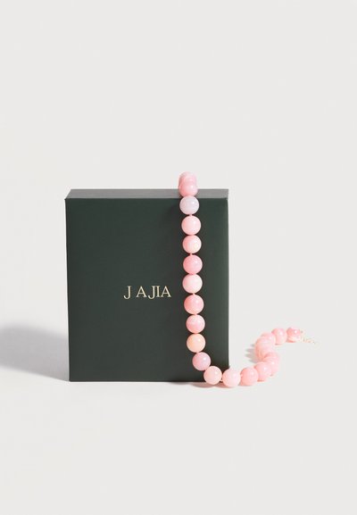 Boîte vert foncé avec texte doré "J AJIA", drapée d'un collier de perles rose avec des perles rondes et brillantes.