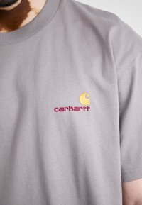 Gray short-sleeve t-shirt made of cotton. Features embroidered yellow and maroon "carhartt" logo on the left chest area.