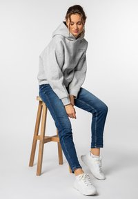 Hellgraue, oversized Kapuzenjacke mit einem Logo auf der Vorderseite, blaue, eng anliegende Jeans und weiße Sneakers, sitzt auf einem Holzschemel vor einem schlichten Hintergrund.
